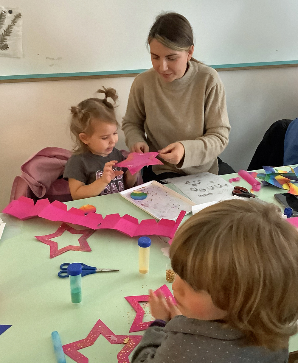 kinder und eine frau sitzen am tisch und schneiden sterne aus bunter pappe aus