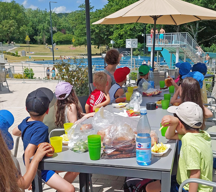 eine-kindergruppe-sitzt-draußen-am-tisch-im-schwimmbad-und-isst-pommes-und-broetchen