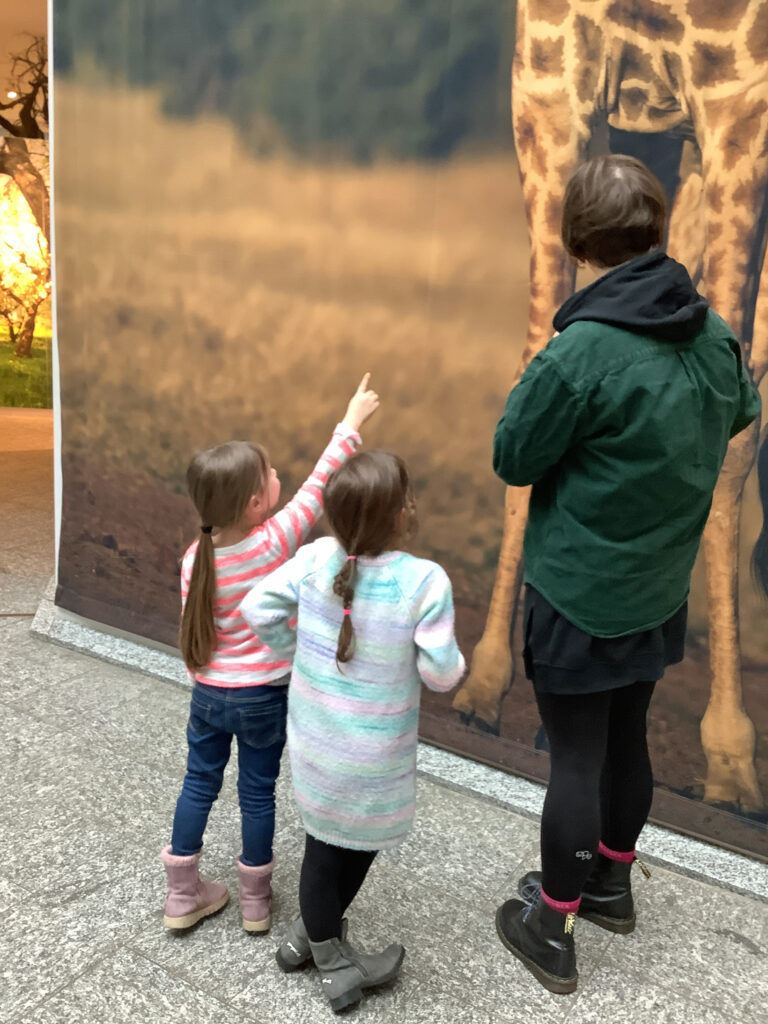 Kinder und eine erwachsene Person betrachten ein riesiges Bild mit einer Giraffe