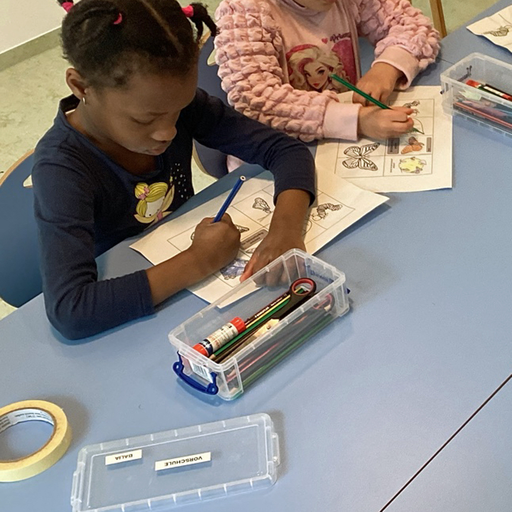 Kinder sitzen am Tisch und malen auf einem Blatt Papier verschiedene Schmetterlinge bunt an