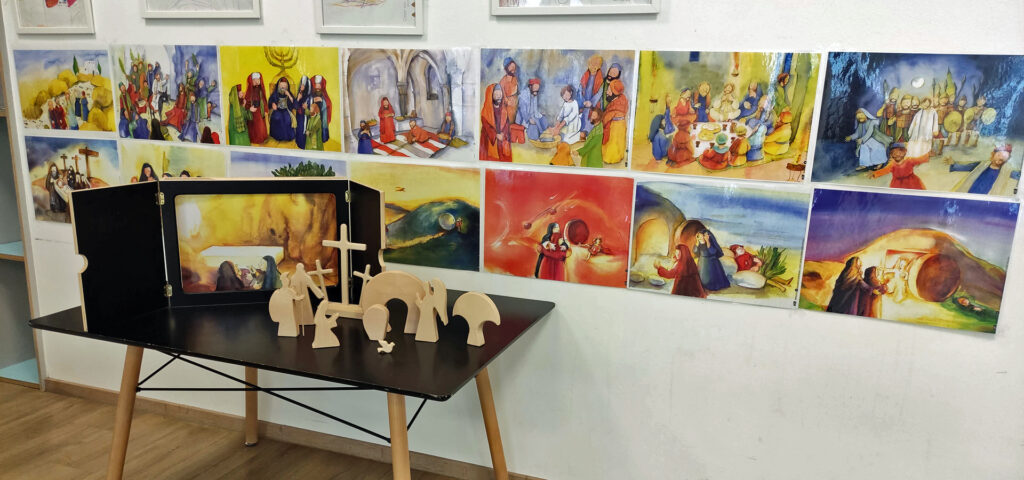 Vor bunten Wandbildern stehen Holzfiguren auf einem Tisch, die das Ostergeschehen symbolisieren