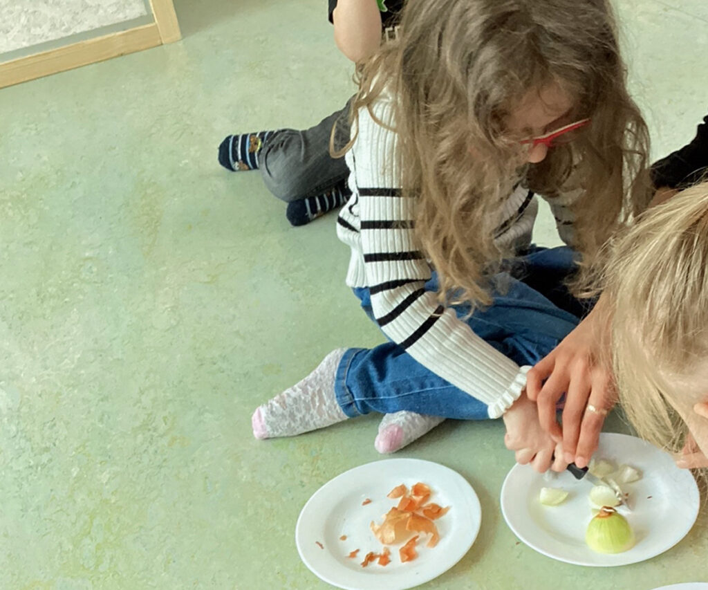 Kinder und Erwachsene hocken auf dem Boden und schneiden eine Zwiebel in Stuecke