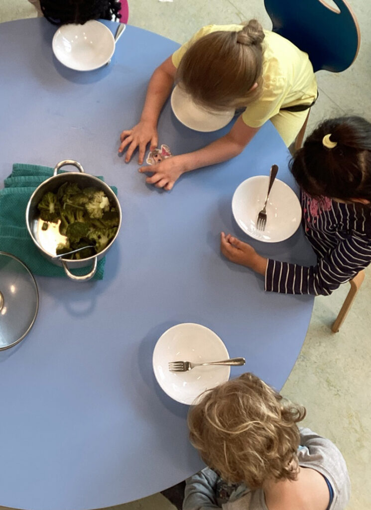 Kinder sitzen am Tisch und in der Mitte steht ein Topf mit gekochtem Brokkoli