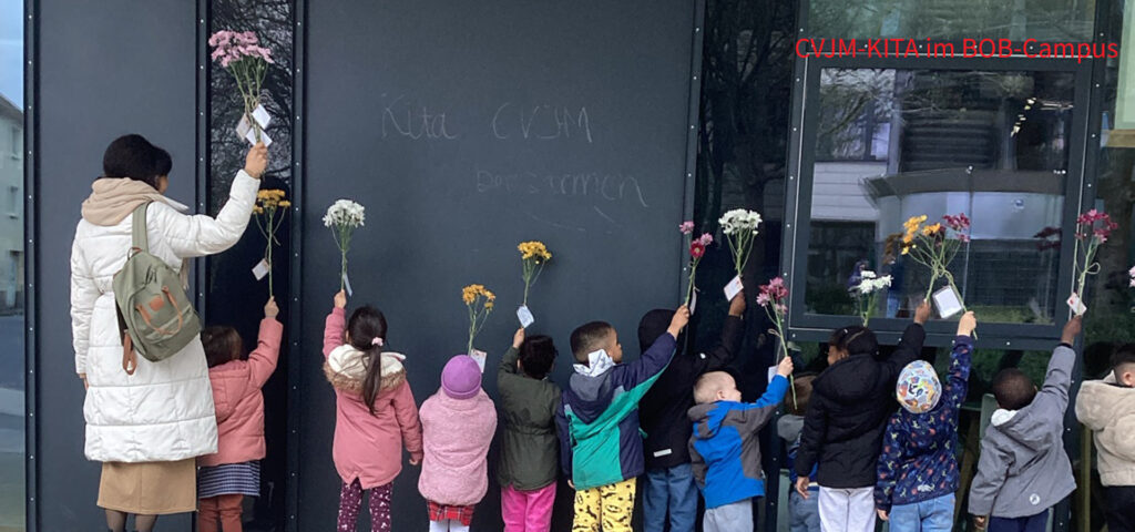 Kinder stehen vor einem Gebaeude und halten Blumen hoch in die Luft