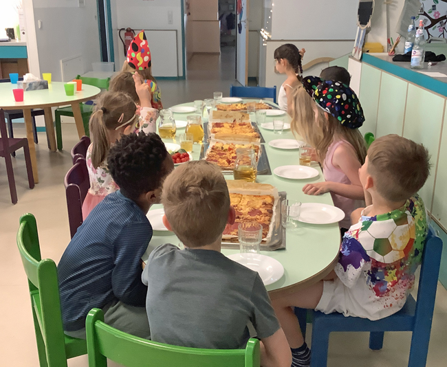 Kinder sitzen an einem Tisch mit Blechen voller selbstgemachter Pizza