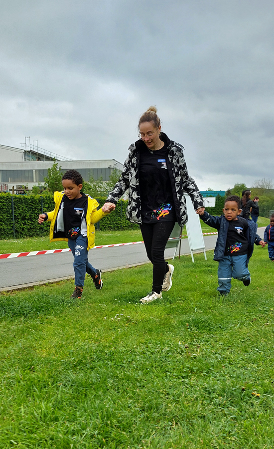 Eine Erzieherin haelt Kinder an den Haenden und alle laufen am Rand einer Wiese entlang