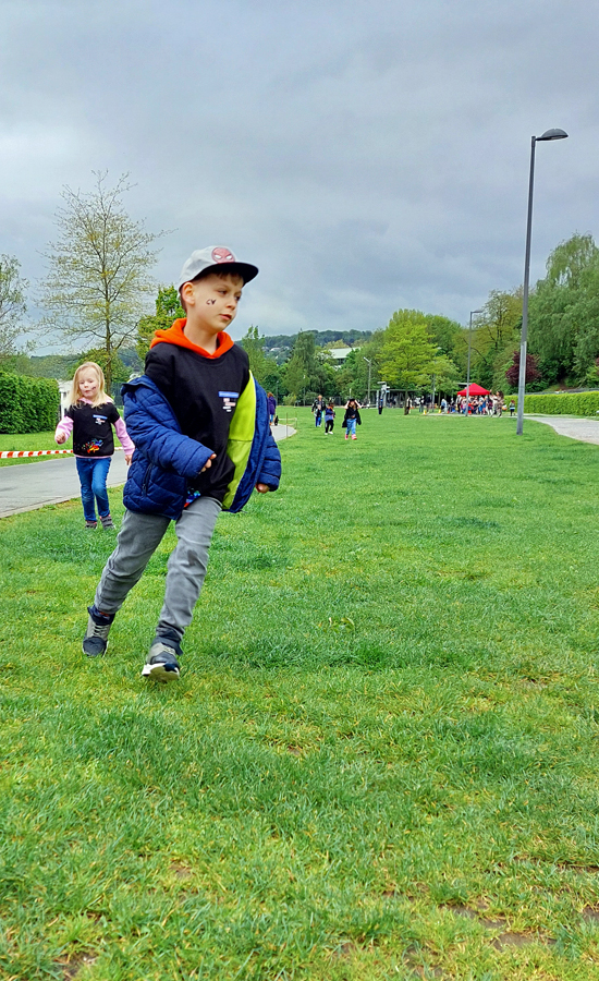 Kinder laufen hintereinander am Rand einer Wiese neben einem rot-weissen Absperrband entlang