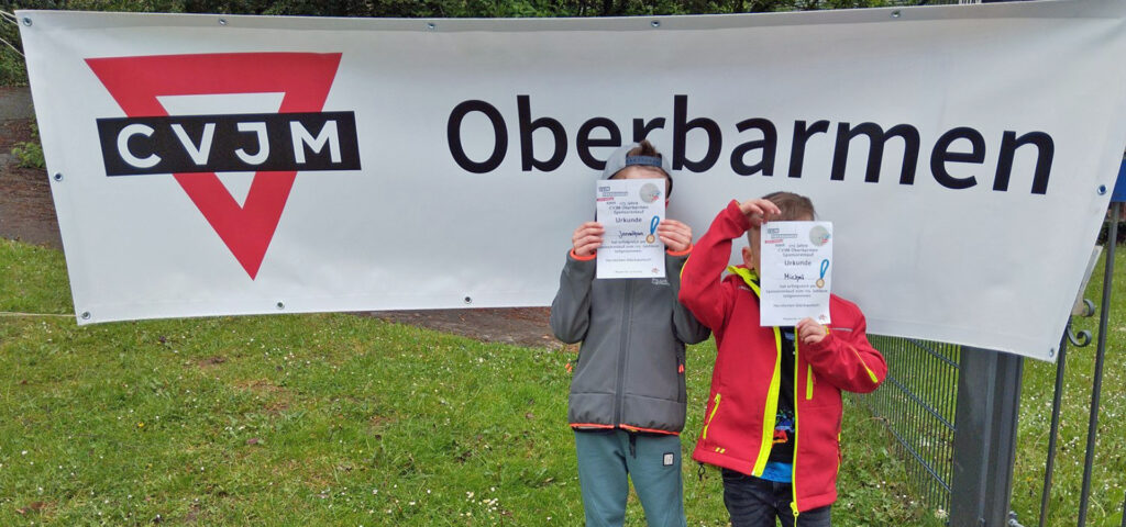 Zwei Kinder halten Urkunden vors Gesicht und stehen vor dem Banner mit der Aufschrift CVJM Oberbarmen