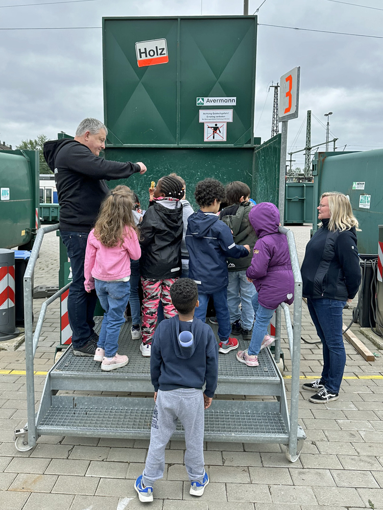 Ein Mitarbeiter des Abfallwirtschaftsbetriebes zeigt Kindern einen grossen Behaelter mit Holzresten