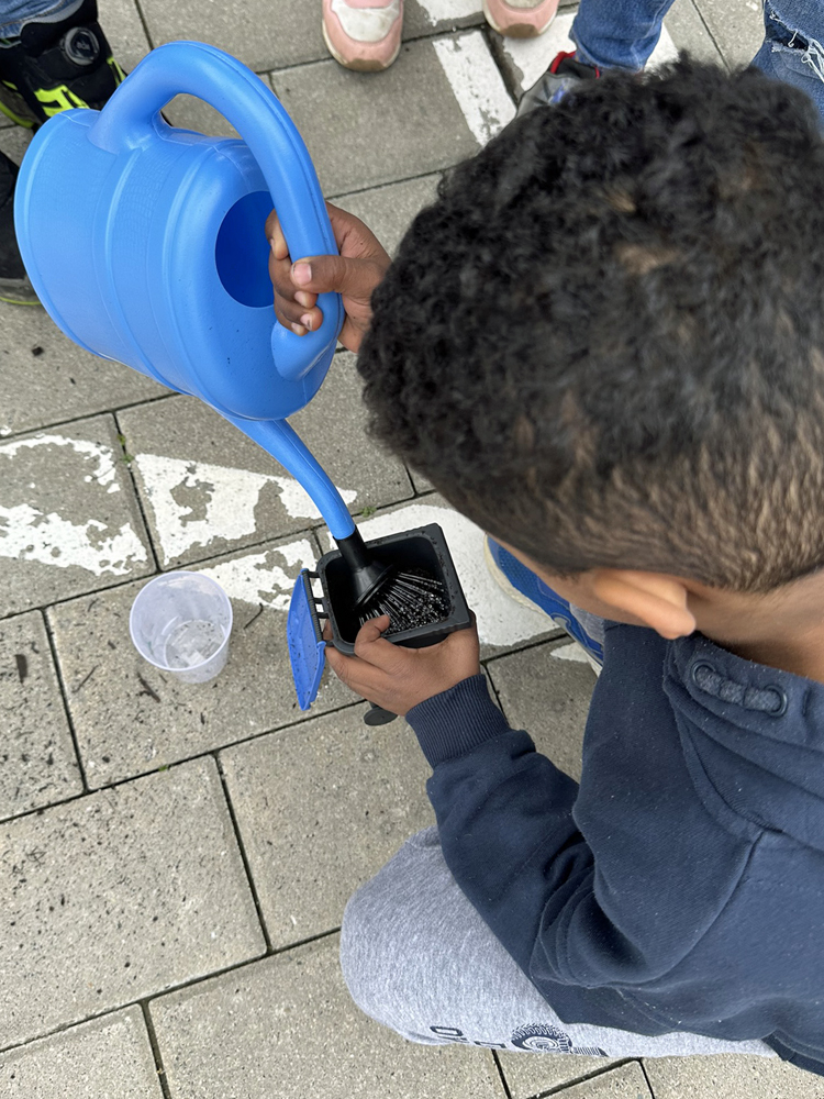 Ein Kind giesst Wasser in eine mit Erde gefuellte Mini-Abfalltonne