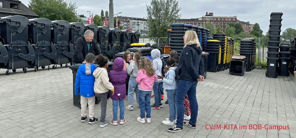 Kinder stehen zwischen vielen neuen Muellbehaeltern und hoeren einem Erwachsenen zu