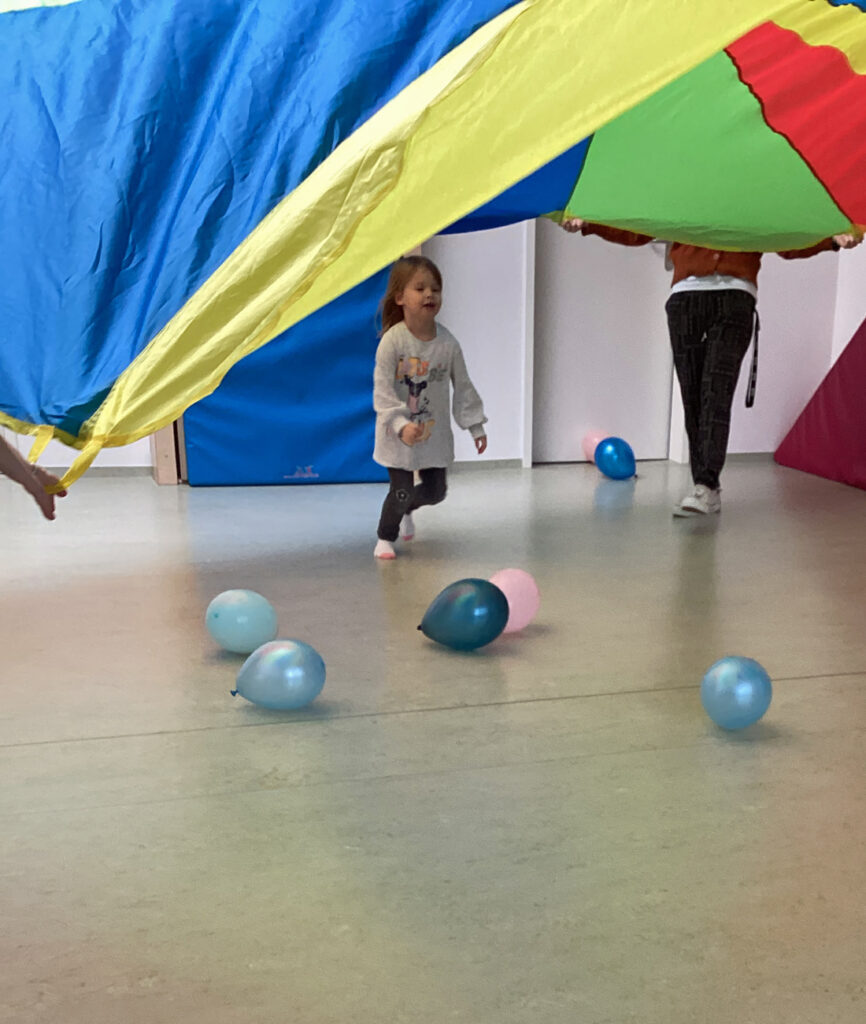 Ein Kind laeuft unter einem bunten Schwungtuch her und auf dem Boden liegen Luftballons