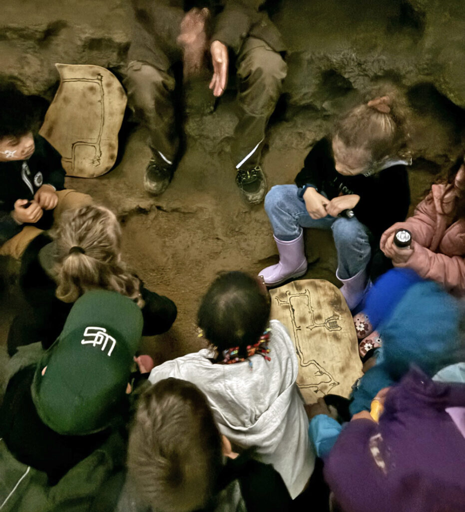 Kinder hocken auf dem Boden einer Hoehle und schauen sich eine Schatzkarte an