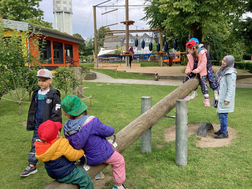 Kinder spielen auf einer großen Wippe aus Holz und eine Erzieherin unterstuetzt sie