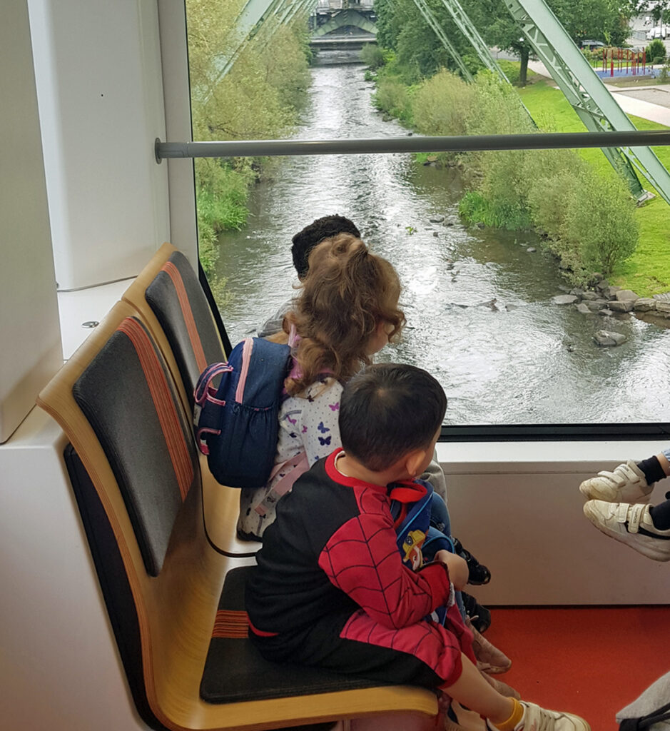 Kinder sitzen auf einer Bank in der Schwebebahn und schauen raus auf die Wupper