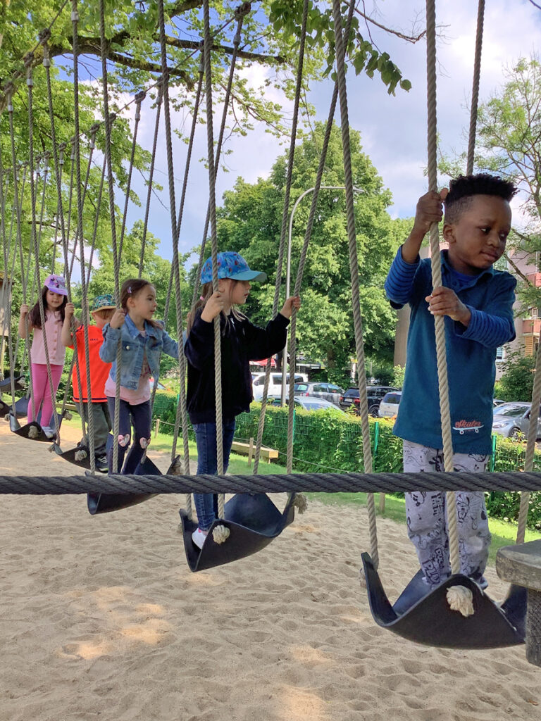 Fuenf Kinder schaukeln und balancieren hintereinander auf einem Spielgeraet aus Seilen