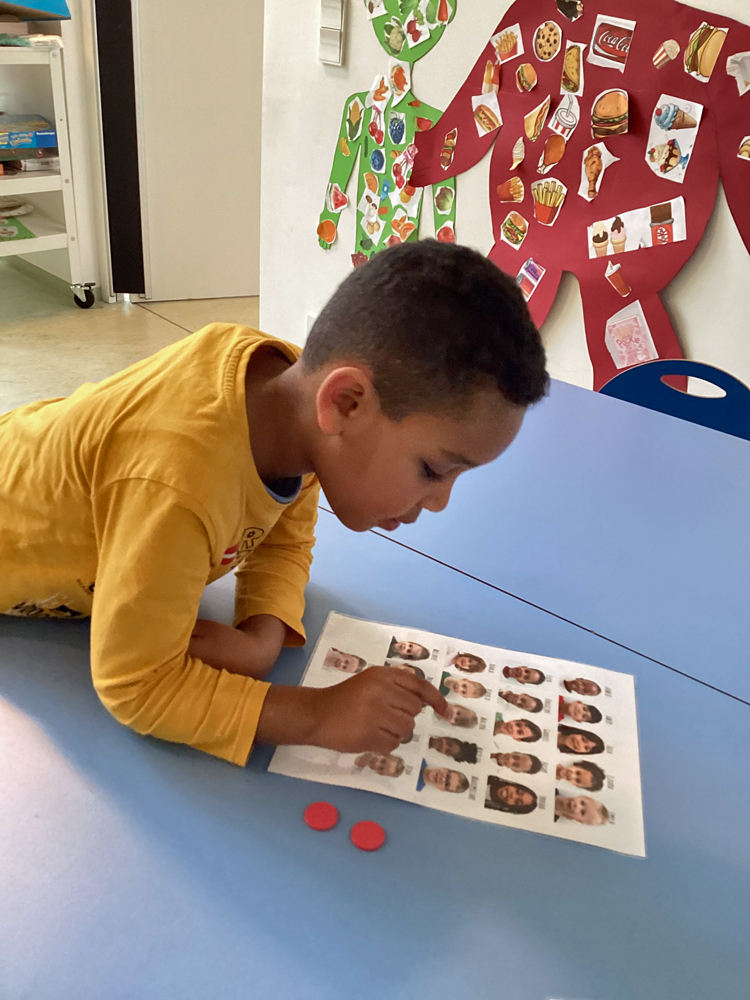 Ein Kind beugt sich ueber einen Tisch und schaut einen Wahlzettel mit Portraitfotos von Kindern an