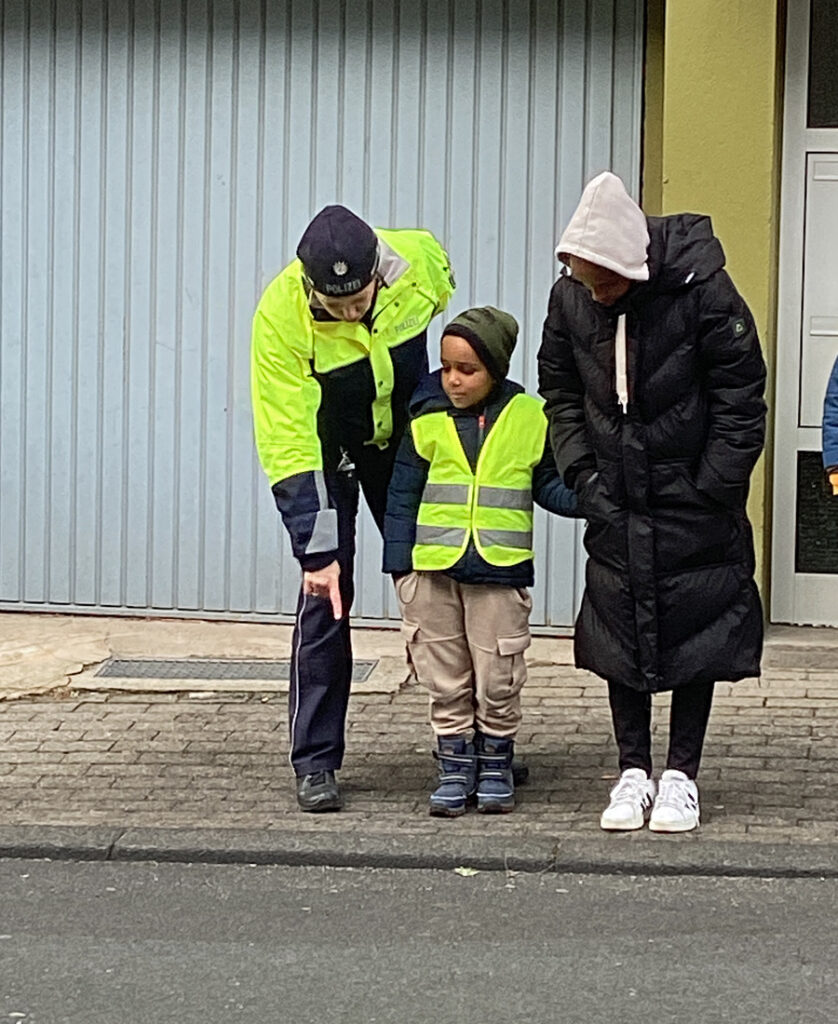 Eine Polizistin steht mit einem Kind und einer Erwachsenen am Strassenrand und zeigt auf den Bordstein