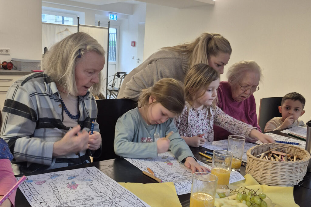 Kinder und Seniorinnen sitzen am Tisch und malen Bilder aus