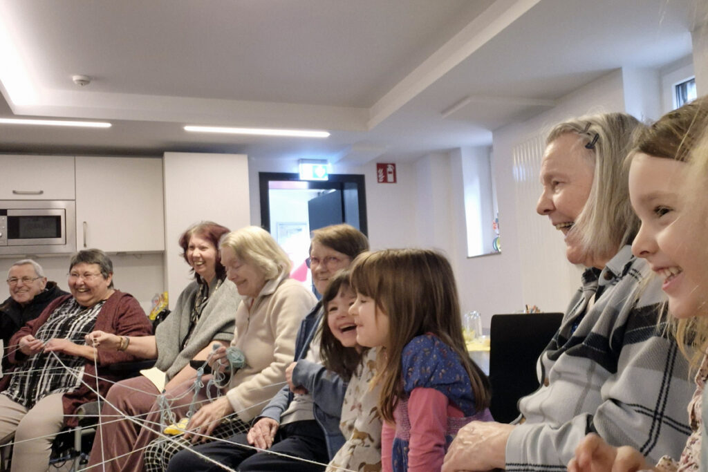 Kinder und Seniorinnen sitzen auf Stuehlen im Kreis und halten Wollfaeden in der Hand und lachen