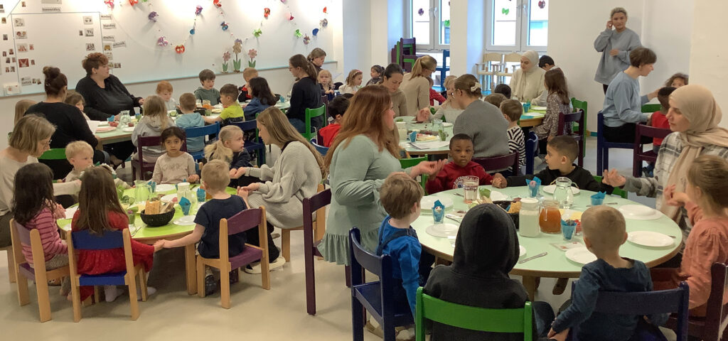 kinder und erwachsene sitzen an bunten tischen und fruehstuecken