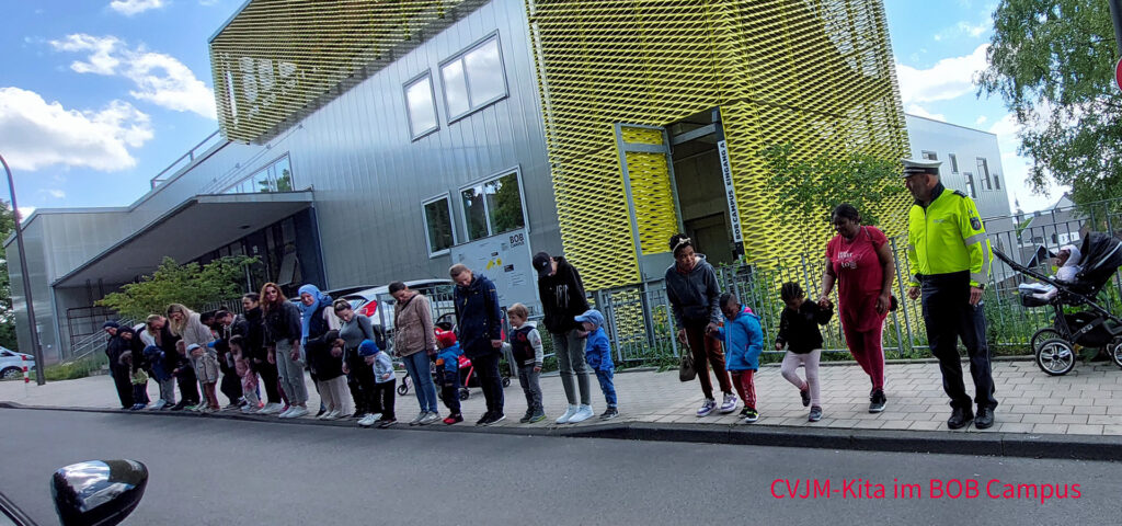 Kinder, Eltern und ein Verkehrspolizist stehen am Strassenrand und schauen nach Fahrzeugen