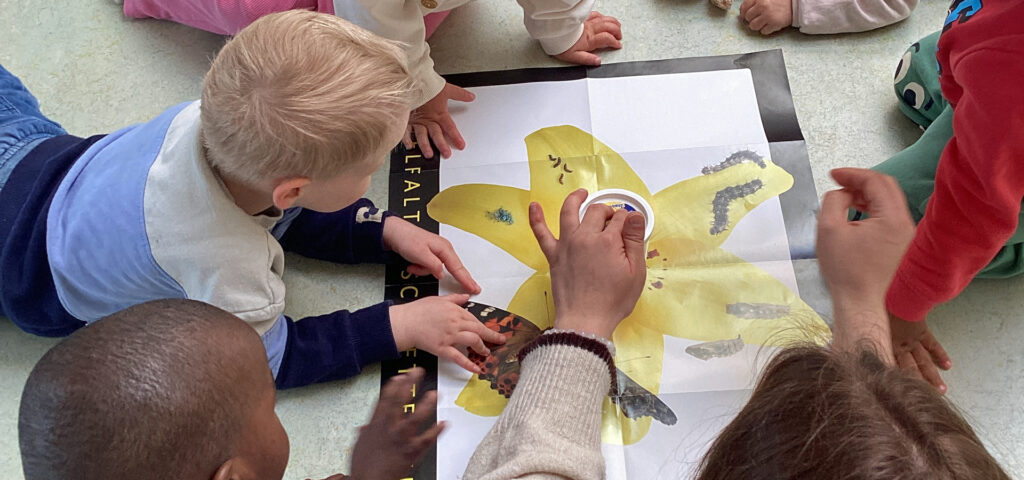 Kinder liegen im Raum baeuchlings um ein Plakat herum auf dem Raupen zu sehen sind