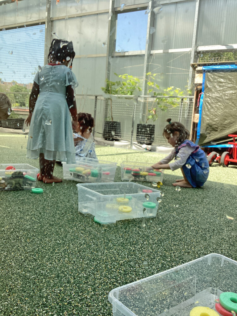 Kinder hocken neben Plastikkisten die mit Wasser gefuellt sind und spielen