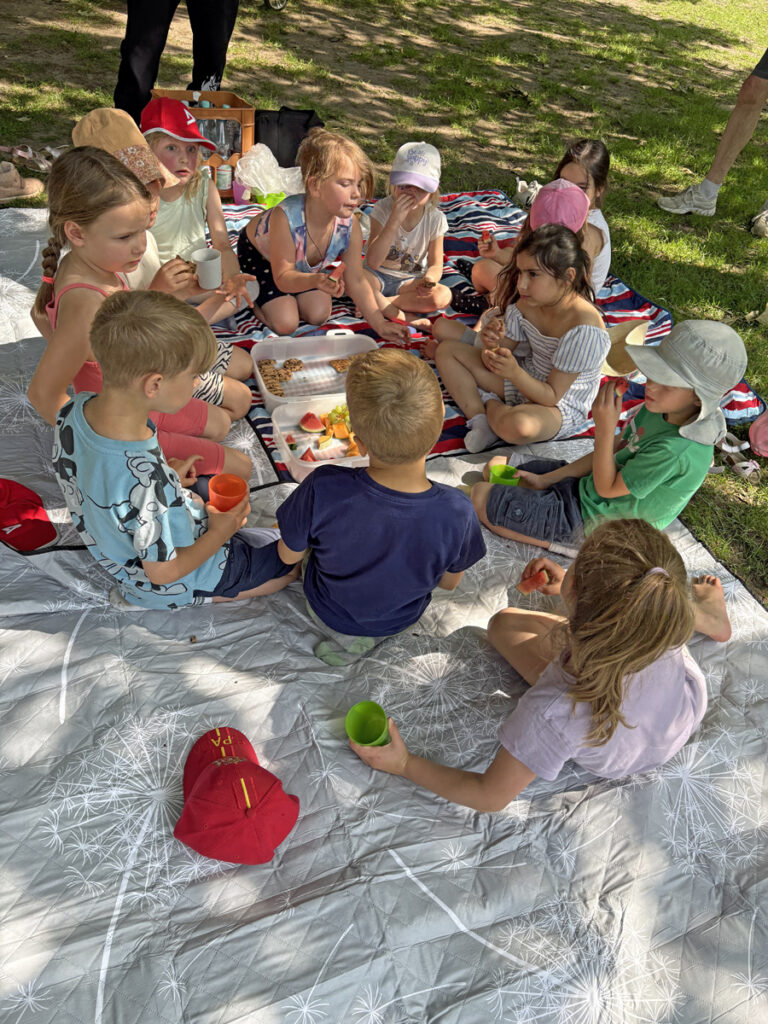 Zwoelf Kinder sitzen auf einer Picknickdecke im Schatten und essen