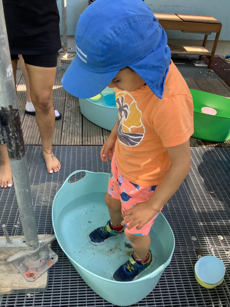 Ein Kind steht draussen mit Schuhen in einem Wasserbottich