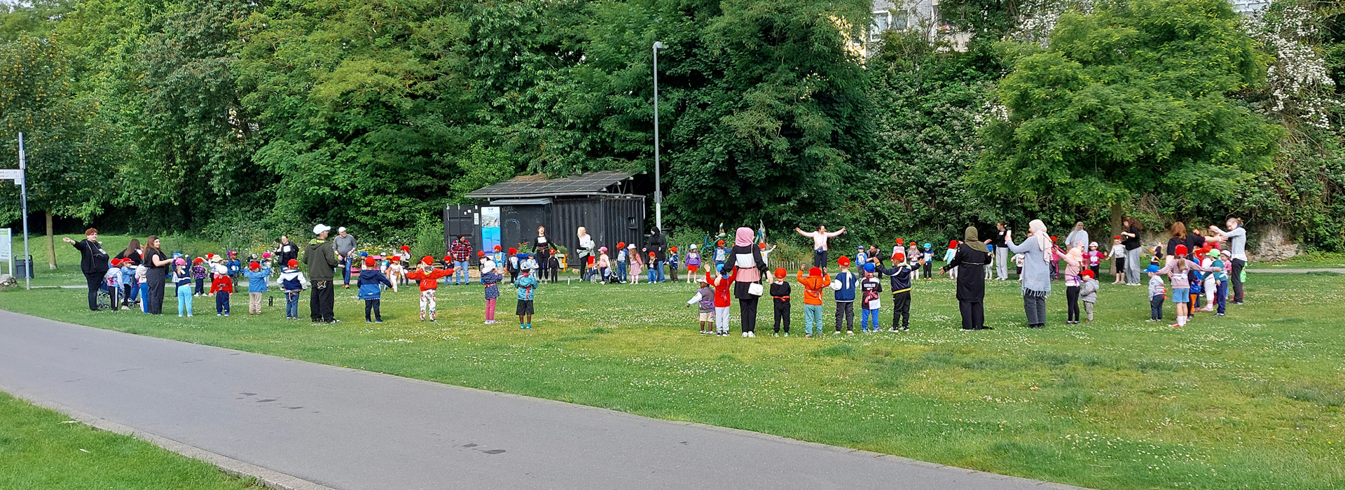 Kinder und Erwachsene stehen im Kreis auf einer Wiese und bewegen die Arme