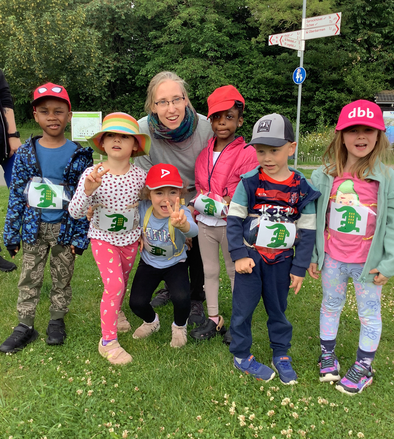 Kinder in Sportkleidung und mit roten Kappen draussen auf einer Wiese schauen mit einer Erzieherin gemeinsam in die Kamera