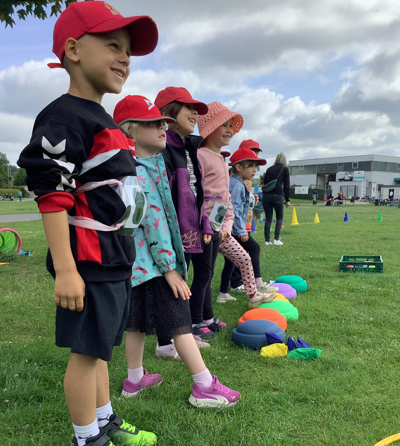 Kinder stehen draussen auf einer Wiese hinter bunten Sportgeraeten und lachen