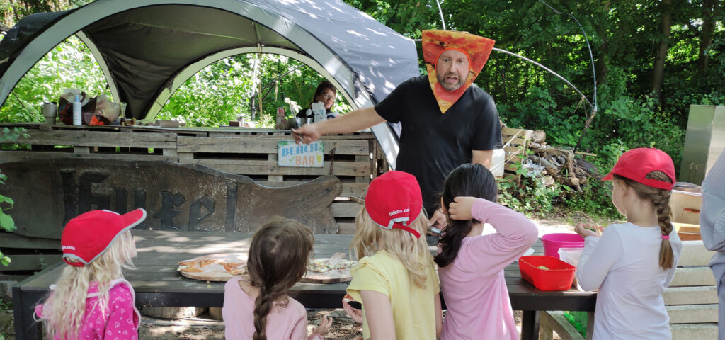 Ein Mann mit Pizza-Verkleidung steht an einer Picknick-Station im Wald vor Kindern und erklaert ihnen etwas