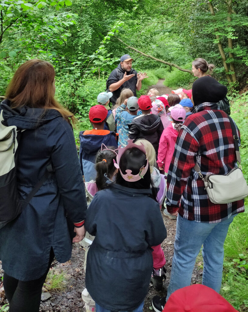 Kinder und Erzieherinnen hoeren einem Mann zu der ihnen im Wald etwas erklaert