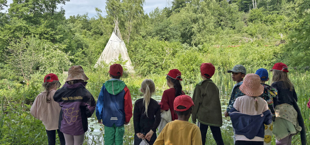 Kinder stehen in der Natur und schauen auf ein Tipi das einige Meter entfernt ist