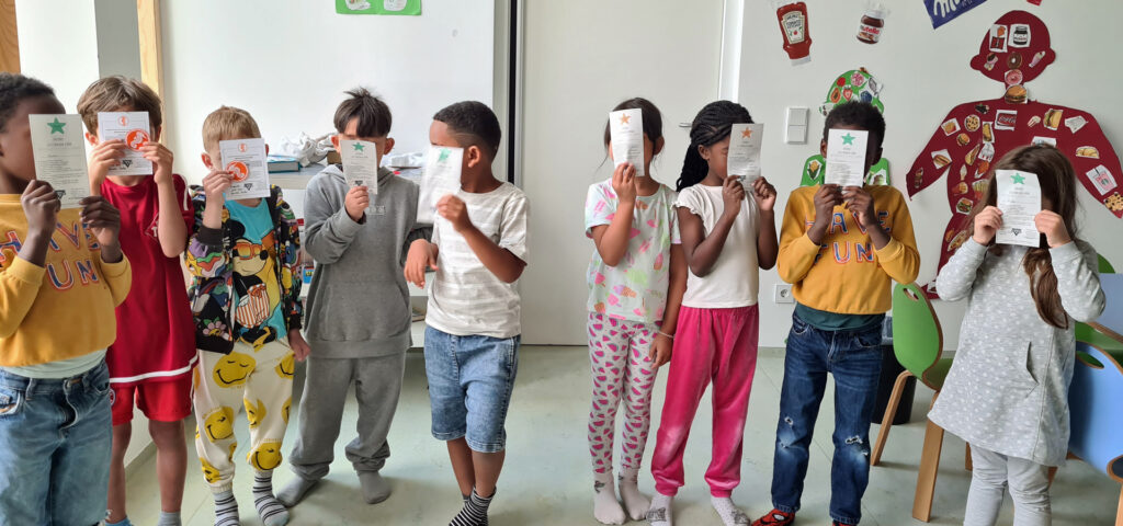 Kinder stehen im Halbkreis und halten Urkunden ueber Schwimmabzeichen vor ihre Gesichter