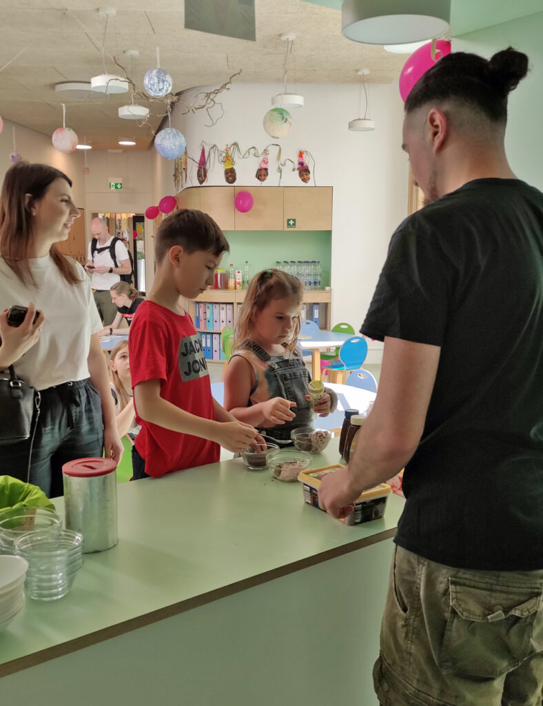 Kinder stehen an einer Theke und fuellen ihre Glasschaelchen mit verschiedenen Eissorten
