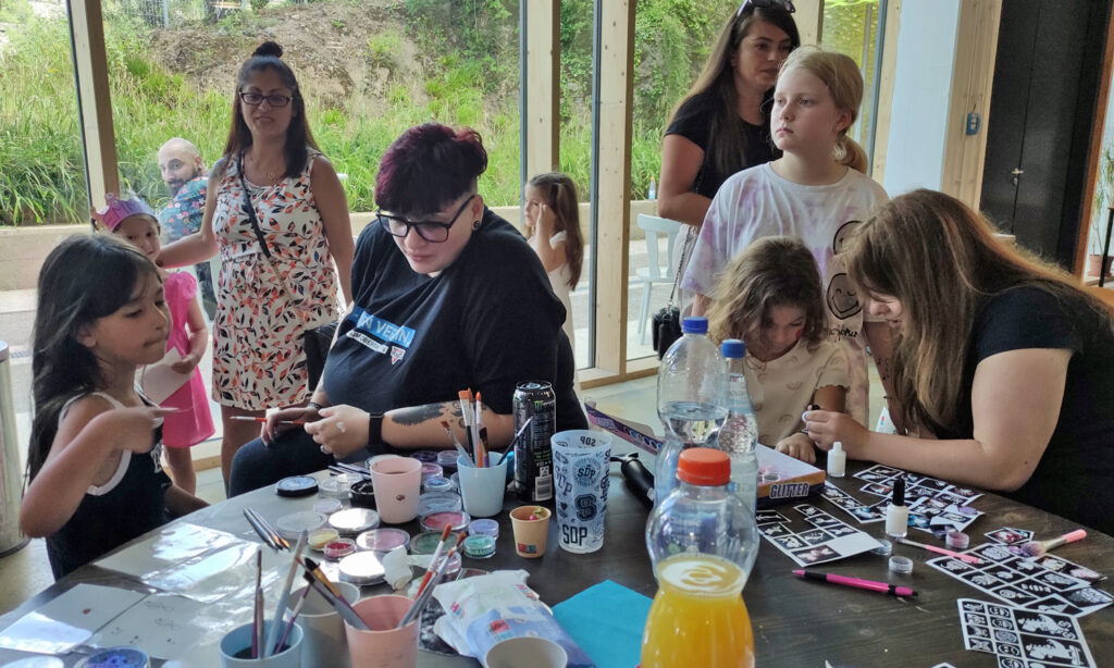 Auf einem Tisch liegen bunte Schminkutensilien plus Tattoos und Erwachsene schminken die Kinder
