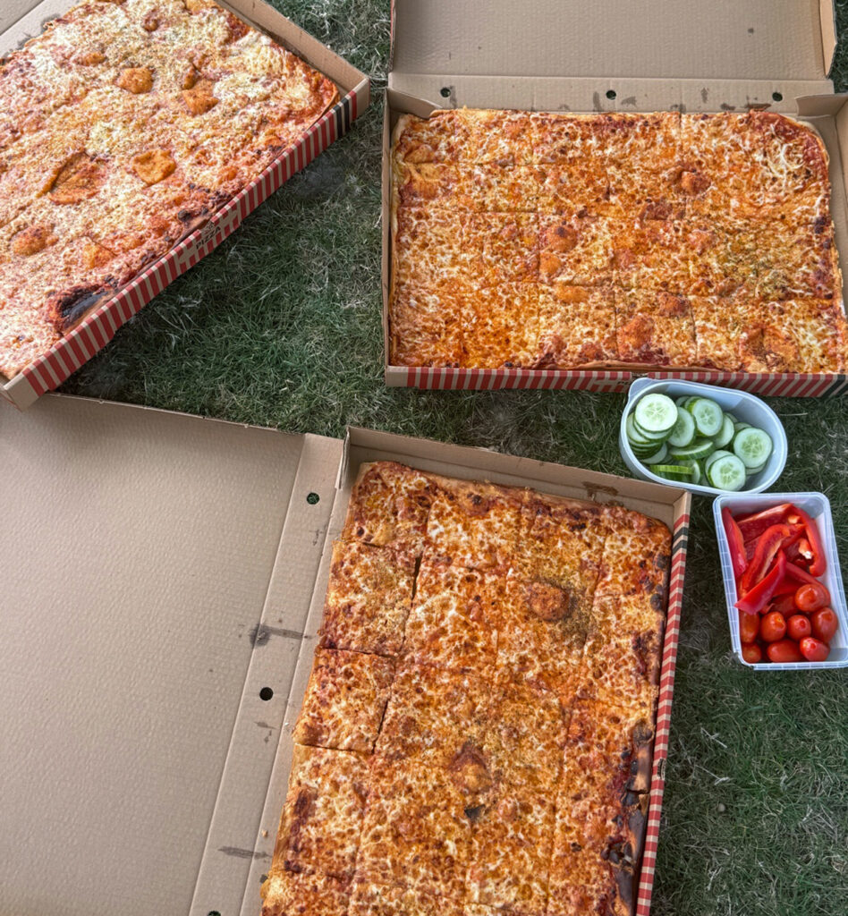 In drei Kartons befindet sich Pizza, daneben stehen Schaelchen mit rohem Gemüse