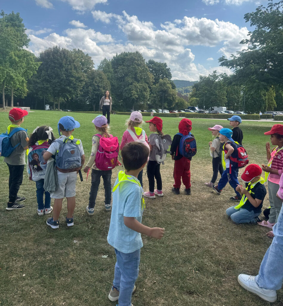 Kinder stehen auf einer Wiese im Park und schauen zu einer Erzieherin