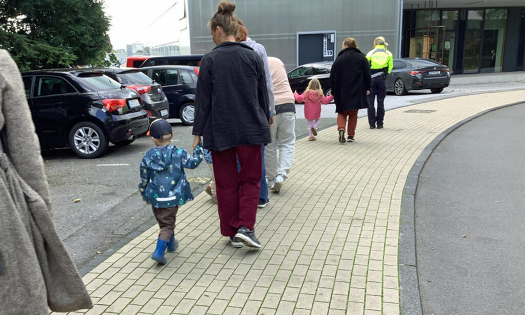 Kinder gehen an der Hand eines Elternteils hinter einer Polizistin auf dem Gehweg und sie befinden sich an der von Strassse abgewandten Seite