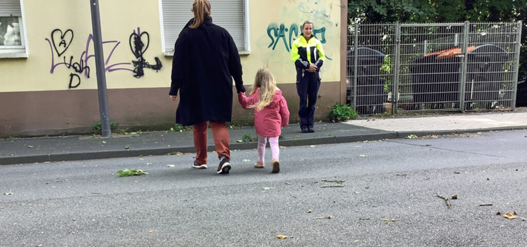 Ein Kind ueberquert an der Hand der Mutter eine Strasse und eine Polizistin schaut zu