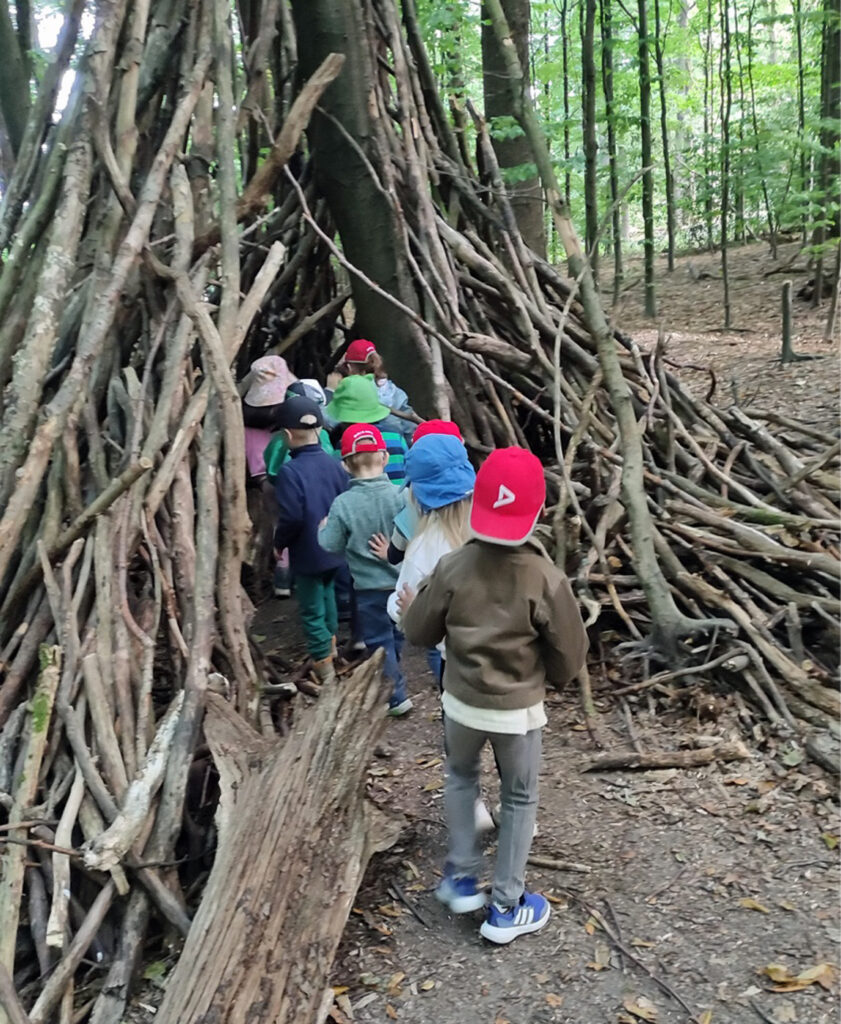 Kinder gehen im Wald in ein aus dicken Aesten lose zusammengebautes Tipi