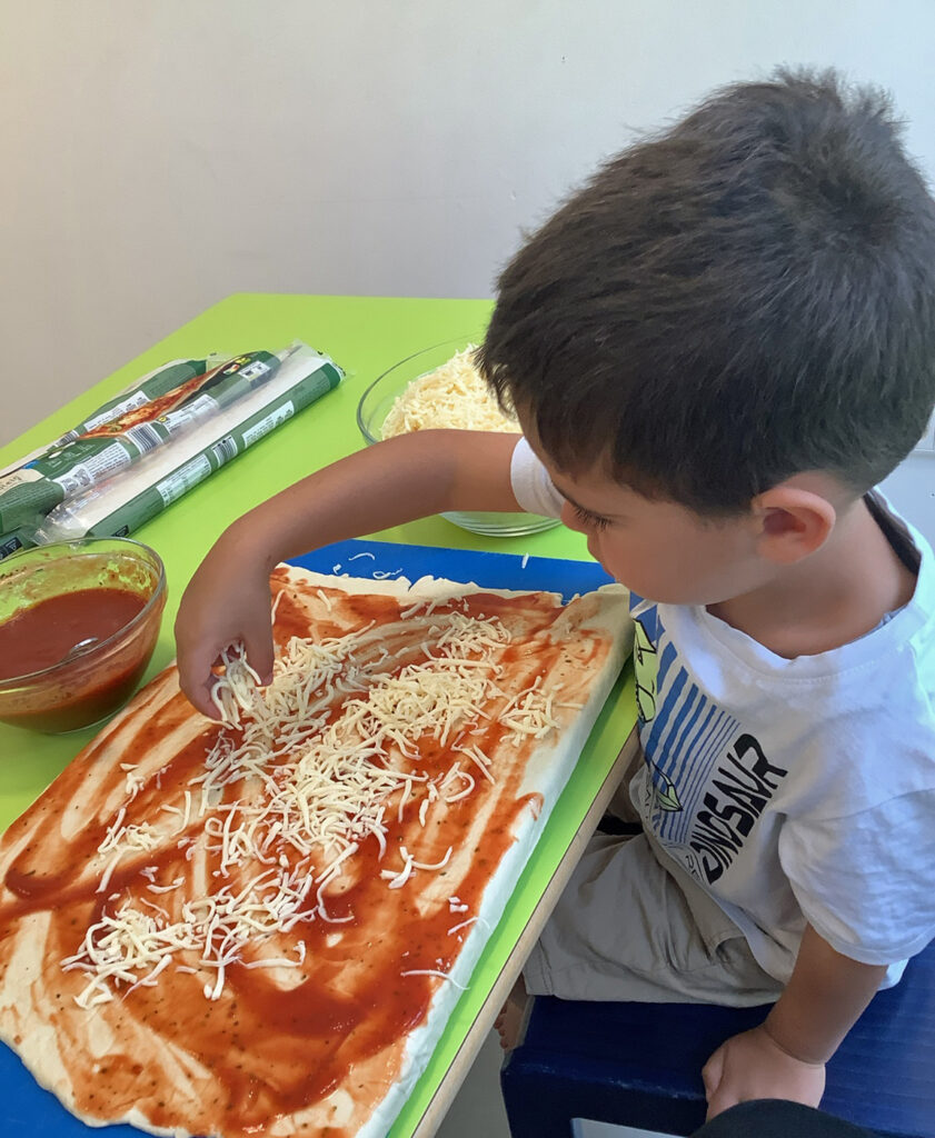 Ein Kind streut Kaese auf Pizzateig und Tomatensauce