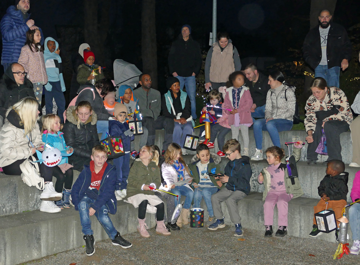 Kinder und Erwachsene halten Laternen in den Haenden und sitzen auf Betonstufen in einem Park
