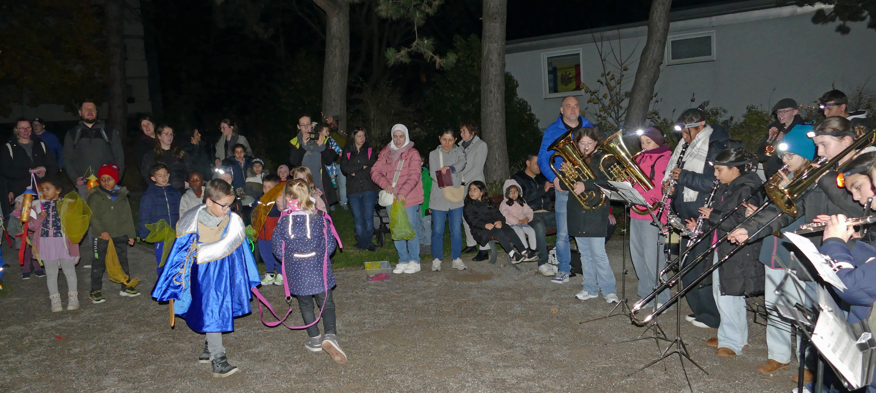 Kinder fuehren draussen in einem Park die Sankt-Martins-Szene auf und Jugendliche mit Blasinstrumenten machen dazu Musik
