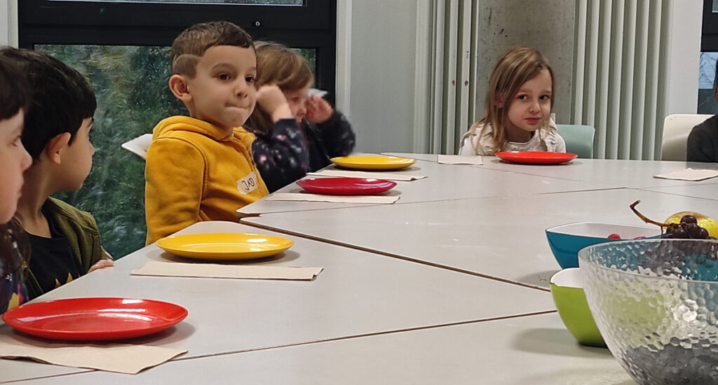 Kinder sitzen am Tisch und haben bunte Teller vor sich und in der Mitte des Tisches stehen Schuesseln mit Obst
