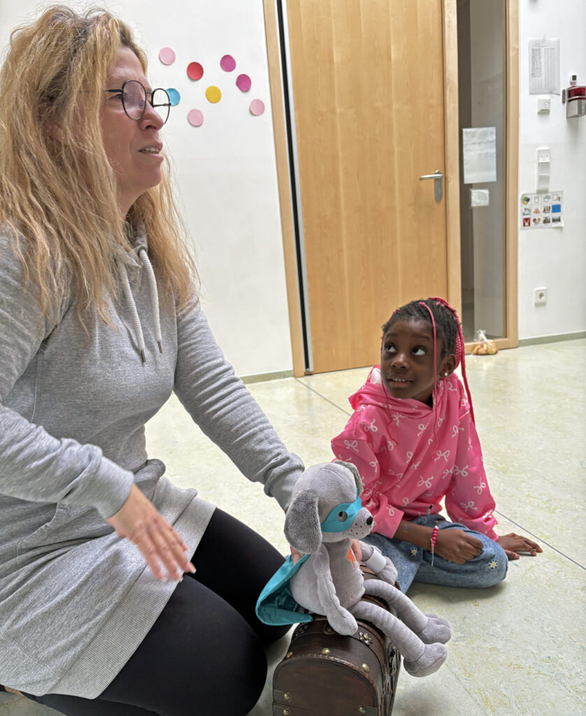 Eine Frau zeigt einen kleinen Stoffhund, der eine Augenmaske und einen Superman-Umhang traegt
