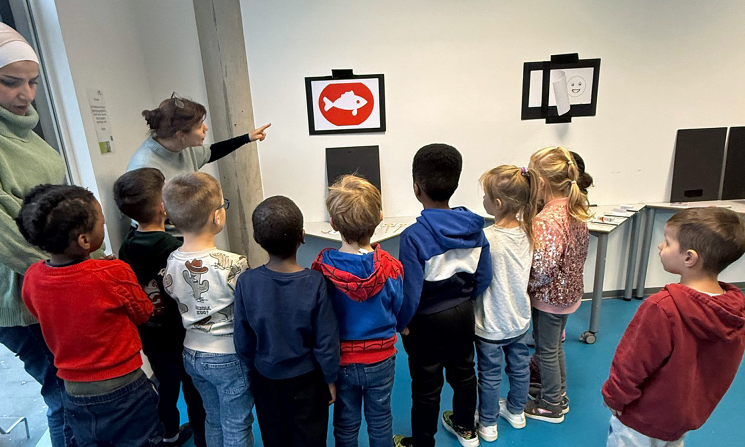 Eine Frau zeigt auf einen weissen Fisch im roten Kreis und eine Gruppe Kinder hoert zu was sie dazu sagt